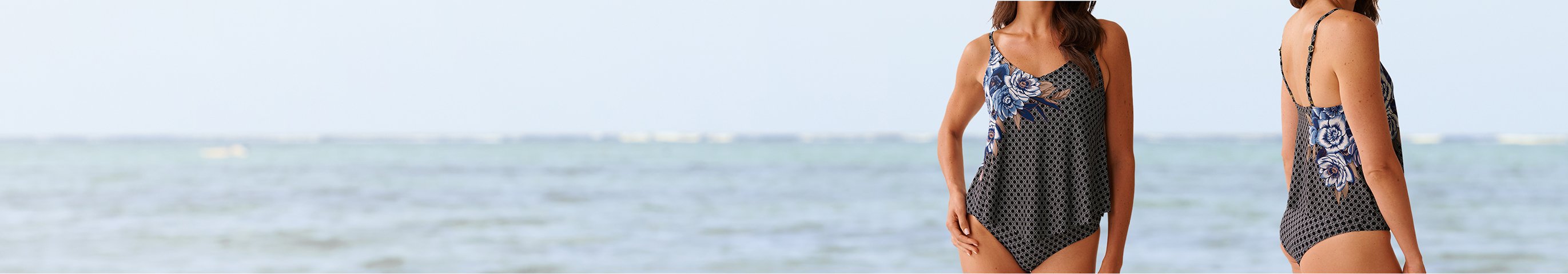 Model seen from front and side in a black tankini with white patterns and a blue flower on the side.